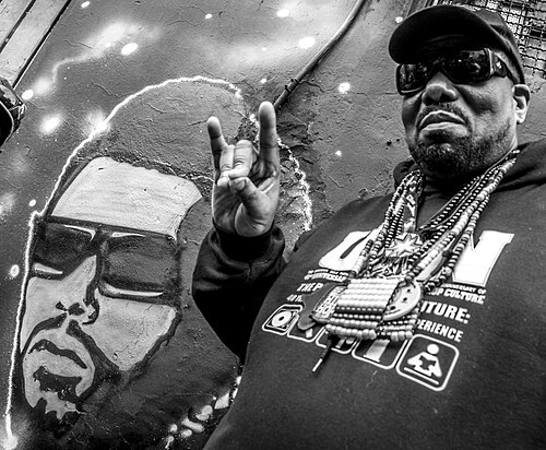 Afrika Bambaataa in a cap and sunglasses, standing next to a mural of himself